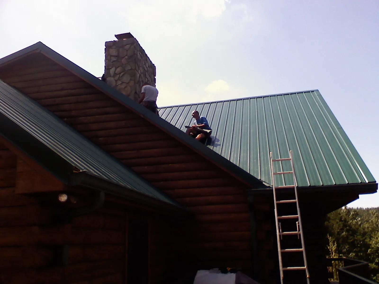 Roofers installing green metal panels beside a stone chimney on a log home in Elkton